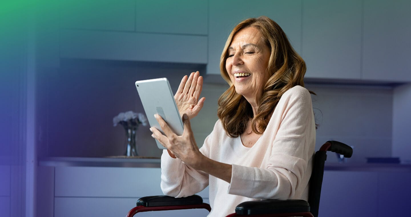 Smiling woman in a light sweater sits in a wheelchair, holding a tablet and waving hello in a kitchen setting.