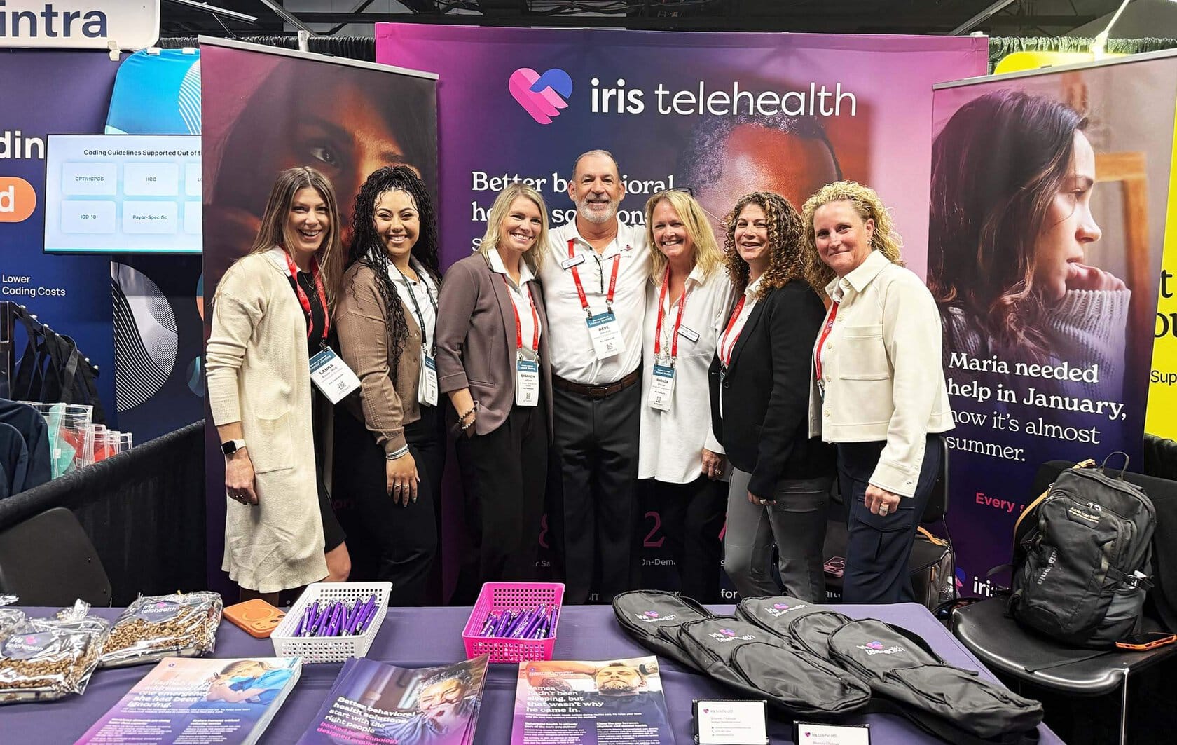 Group of eight people wearing badges posing at a trade show booth for Iris Telehealth in front of purple banners.